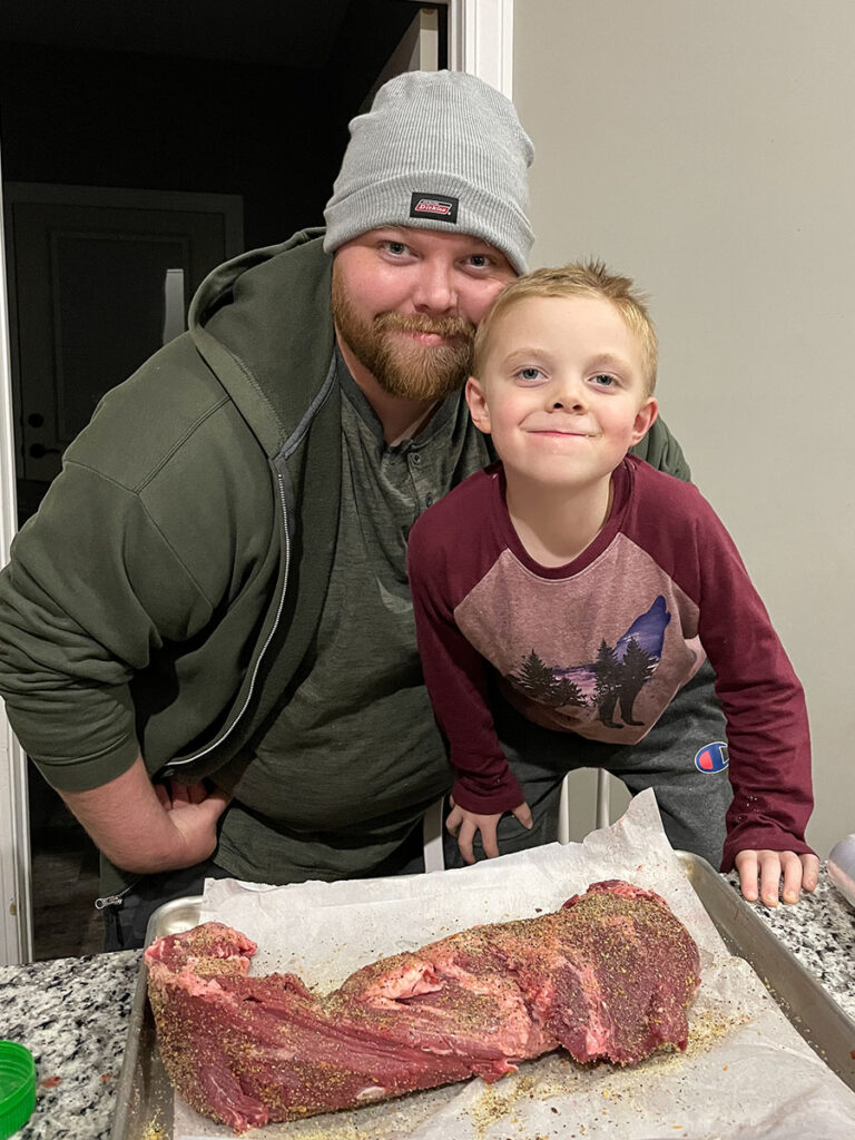 A proud man and a boy smiling while preparing a smoked beef tenderloin.