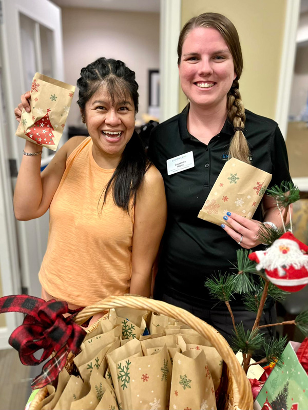 Two team members at a senior living community happily hold up festive treat bags during a Christmas in July celebration, spreading joy and holiday spirit.