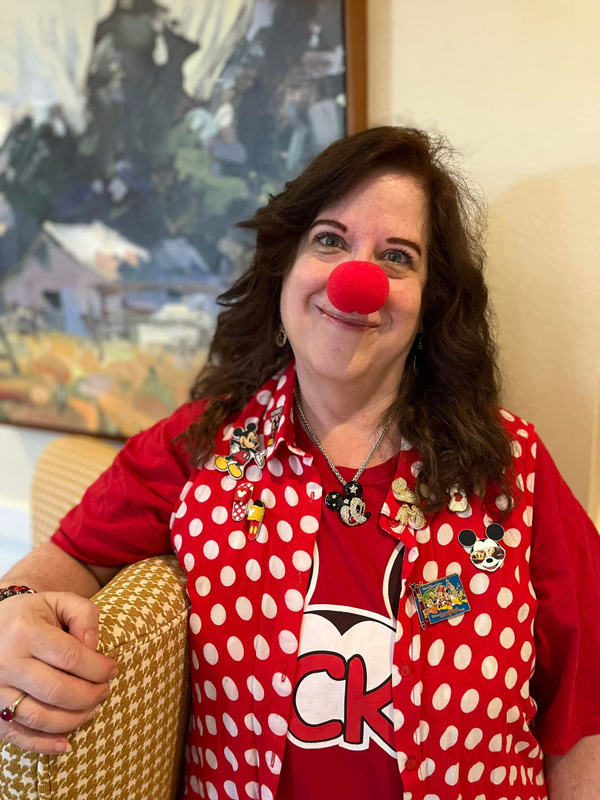 A smiling woman at a senior living community wears a red clown nose and a polka-dot vest adorned with Disney pins, embracing a fun and playful atmosphere.