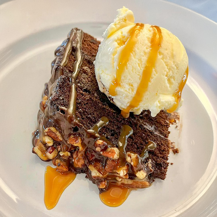 Rich chocolate turtle cake topped with vanilla ice cream and drizzled with caramel sauce, featuring pecans and layers of chocolate for a decadent dessert on a white plate.