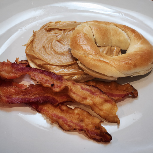 Open-faced bagel topped with peanut butter, served with three crispy bacon strips on a white plate.