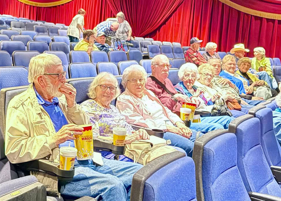 Senior residents seated in a theater, smiling and enjoying popcorn and drinks while waiting for a movie to begin.