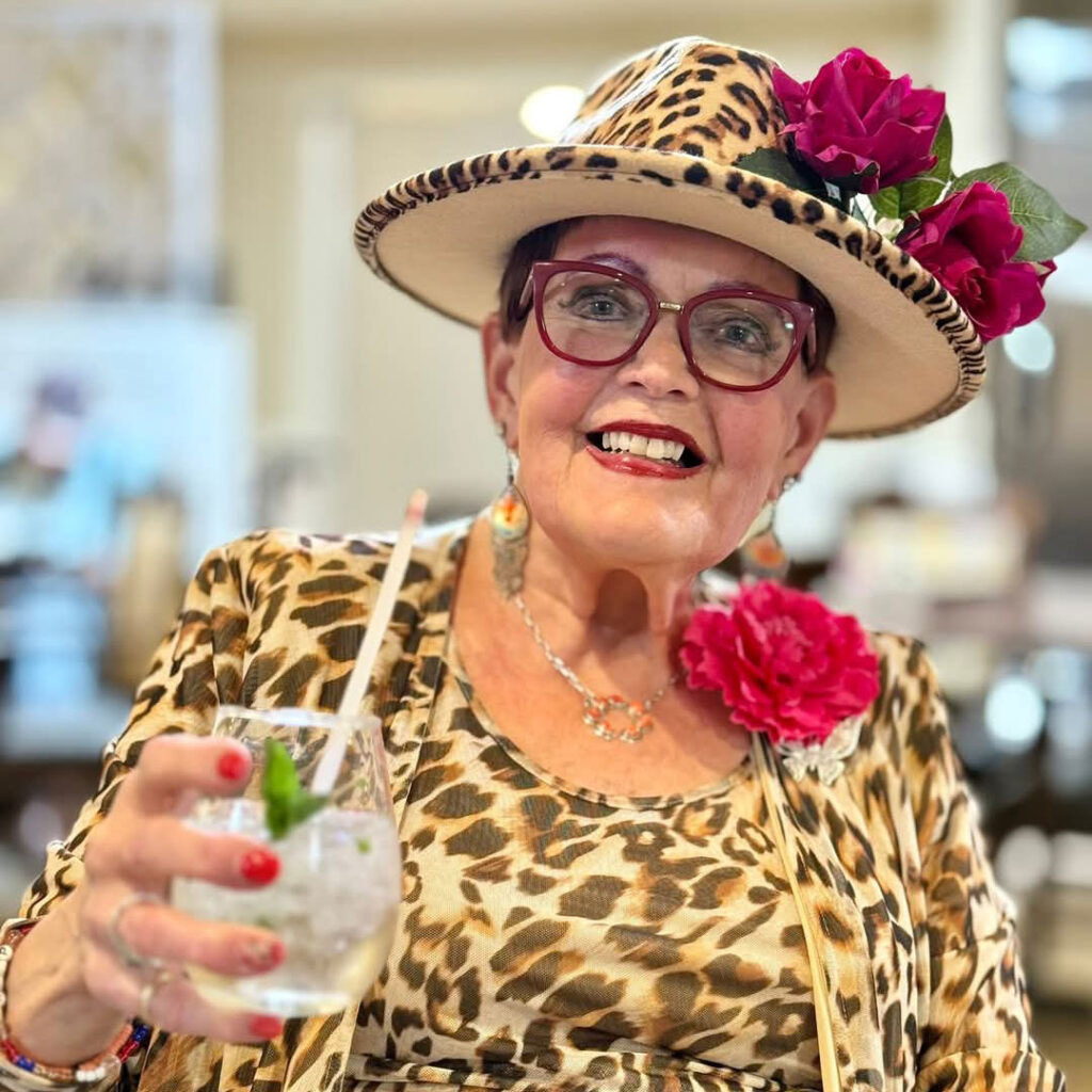 Stylish senior woman wearing a leopard-print dress and hat adorned with red roses, smiling and holding a cocktail with a straw and mint garnish.