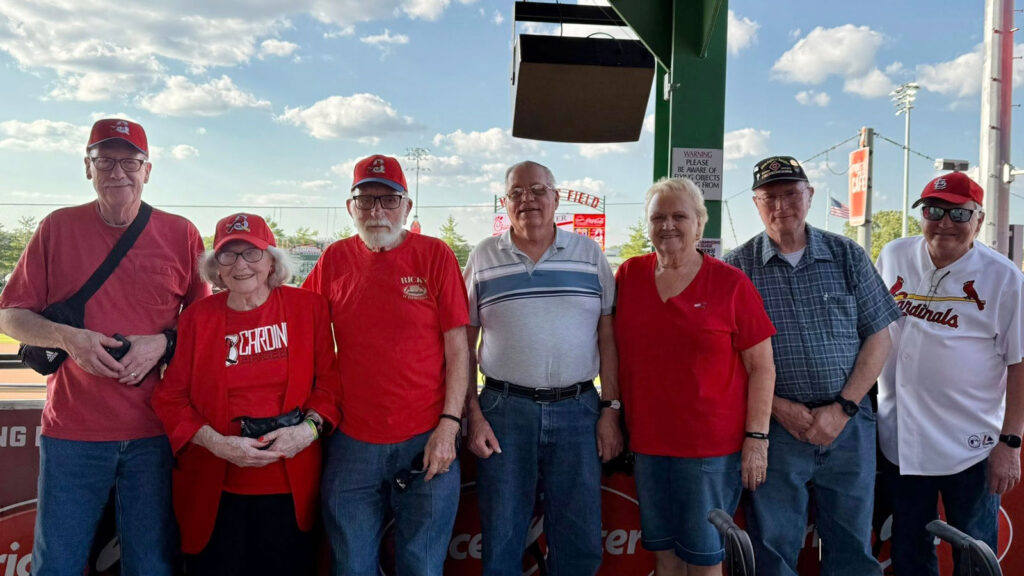A group of senior residents dressed in red and Cardinals gear pose together at a Springfield Cardinals baseball game under a sunny sky.