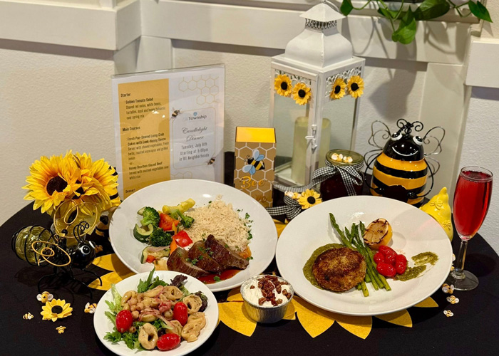 A beautifully arranged table features gourmet dishes including steak, crab cakes, tortellini salad, and dessert, surrounded by bee-themed décor and a honey-inspired menu.