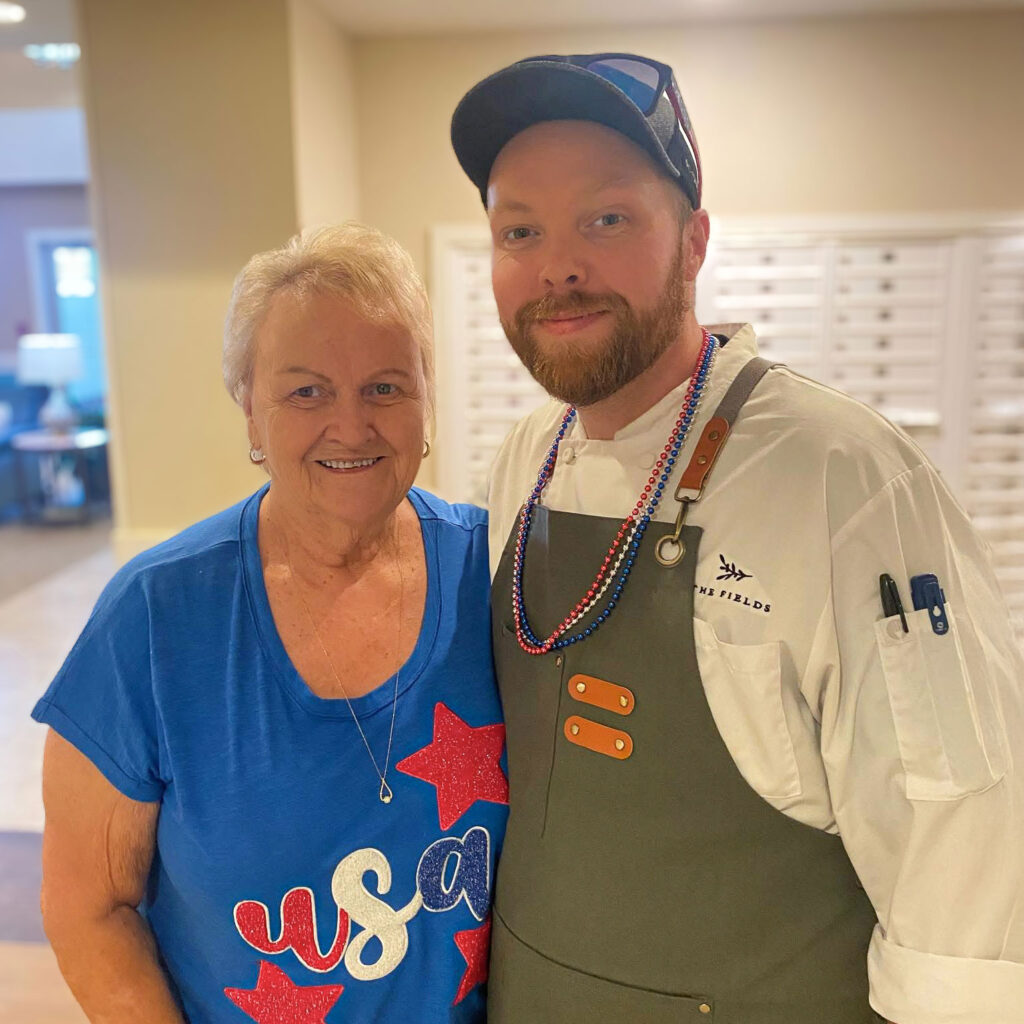 A Culinary Director wearing festive beads smiles with a senior resident dressed in a USA shirt, celebrating community spirit and friendship in a warm setting.
