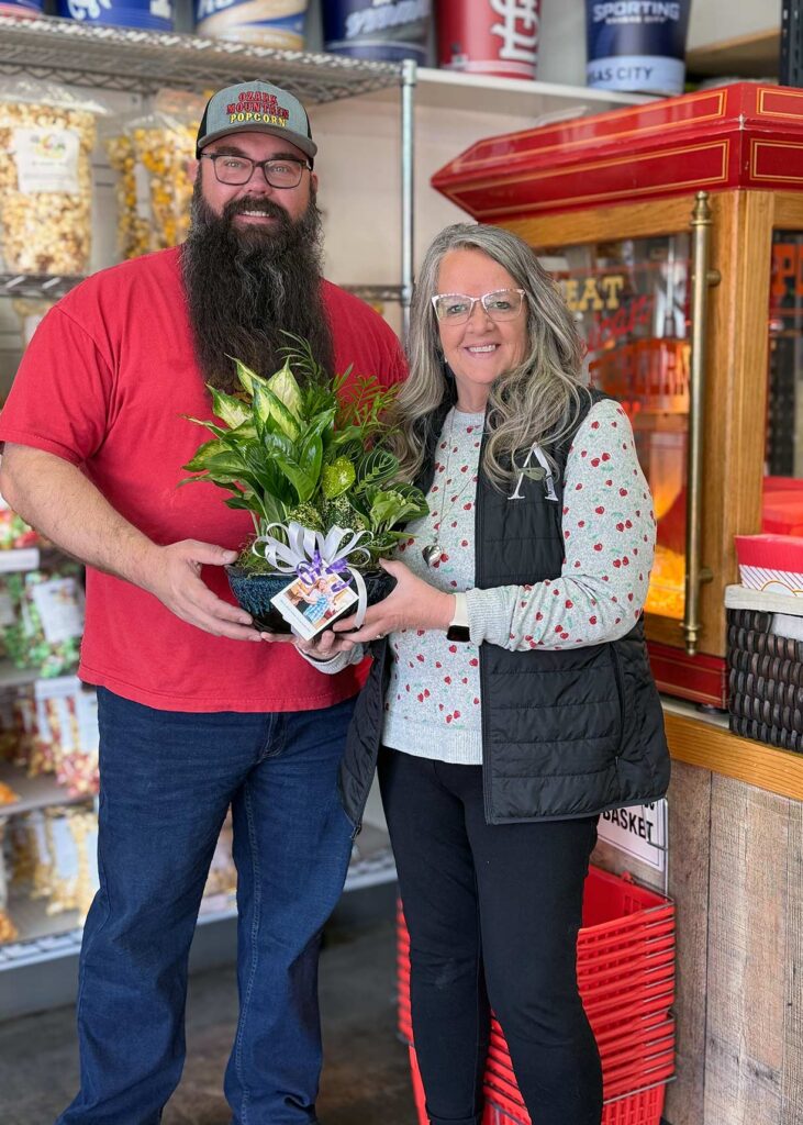 A woman on The Township Senior Living team smiles alongside a local business owner, presenting a beautiful plant.