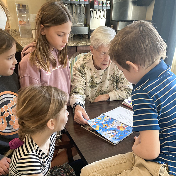 A senior woman reads from a children's book while boys and girls gather around her.