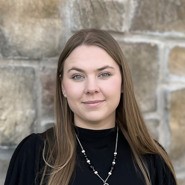 Sierra Jewell, Senior Living Coordinator supporting The Township Senior Living, looking at the camera in a professional headshot with long straight light brown hair, a black top, a pendant necklace, and a stone wall background.
