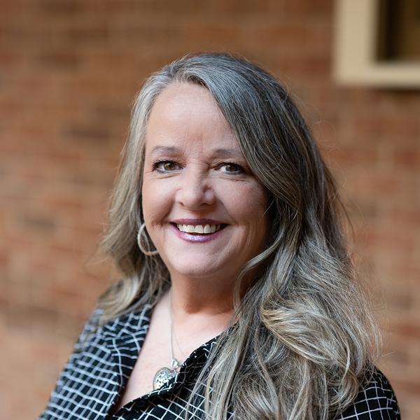 Becky Meagher, Wellness Director at The Township Senior Living, smiling in a professional outdoor headshot with long gray-blonde hair, hoop earrings, a patterned black and white blouse, and a softly blurred brick background.