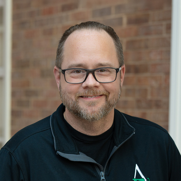Jason Carr, Plant Operations Director at The Township Senior Living, looking at the camera in a professional outdoor headshot with glasses, short hair pulled back, a trimmed beard, a black quarter-zip top, and a softly blurred brick background.