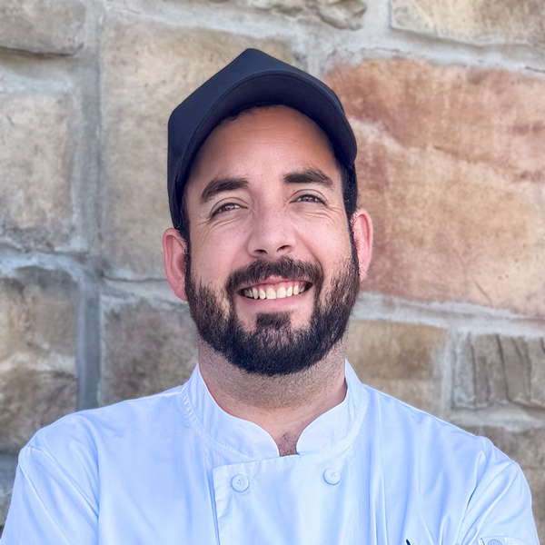 Grant Smith, Culinary Director at The Township Senior Living, smiling in a professional headshot with a black cap, a full dark beard, a white chef coat, and a light stone wall background.