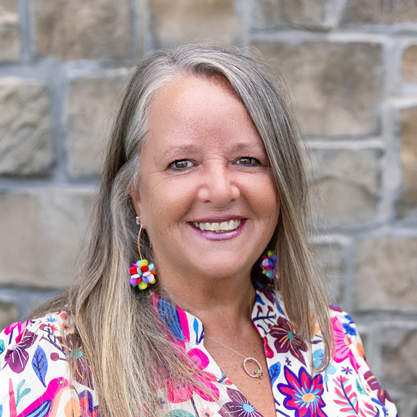 Becky Meagher, Wellness Director at The Township Senior Living, smiling in a professional headshot with long gray-blonde hair, colorful floral earrings, a bright floral blouse, and a stone wall background.