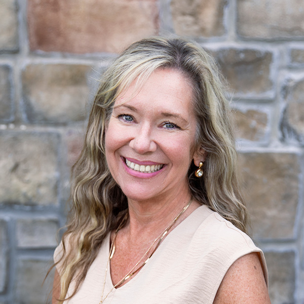 Christy Brashers, Operations Coordinator at The Township Senior Living, smiling in a professional headshot with long wavy blonde hair, pearl drop earrings, a beige sleeveless top, and a stone wall background.