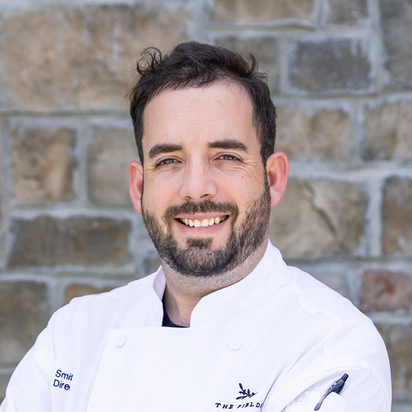 Grant Smith, Culinary Director at The Township Senior Living, smiling in a professional headshot wearing a white chef coat, with short dark hair, a full beard, and a stone wall background.