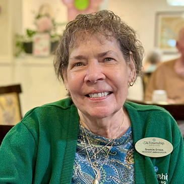 A senior woman smiles at The Township Senior Living, wearing a green cardigan and a gold Resident Ambassador badge.