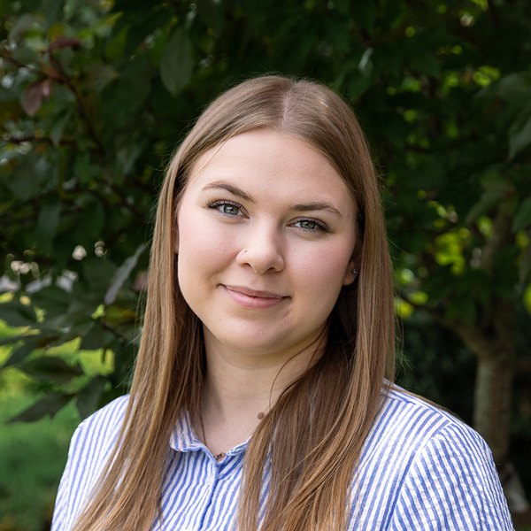 Sierra Jewell, Senior Living Coordinator supporting The Township Senior Living, looking at the camera in a professional outdoor headshot with long straight light brown hair, a blue striped button-up shirt, and a softly blurred green foliage background.